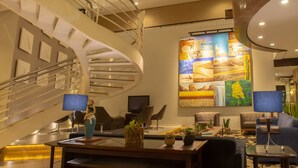 Um lobby moderno com escadaria em espiral, uma grande pintura abstrata e uma área de estar com mesas e cadeiras.