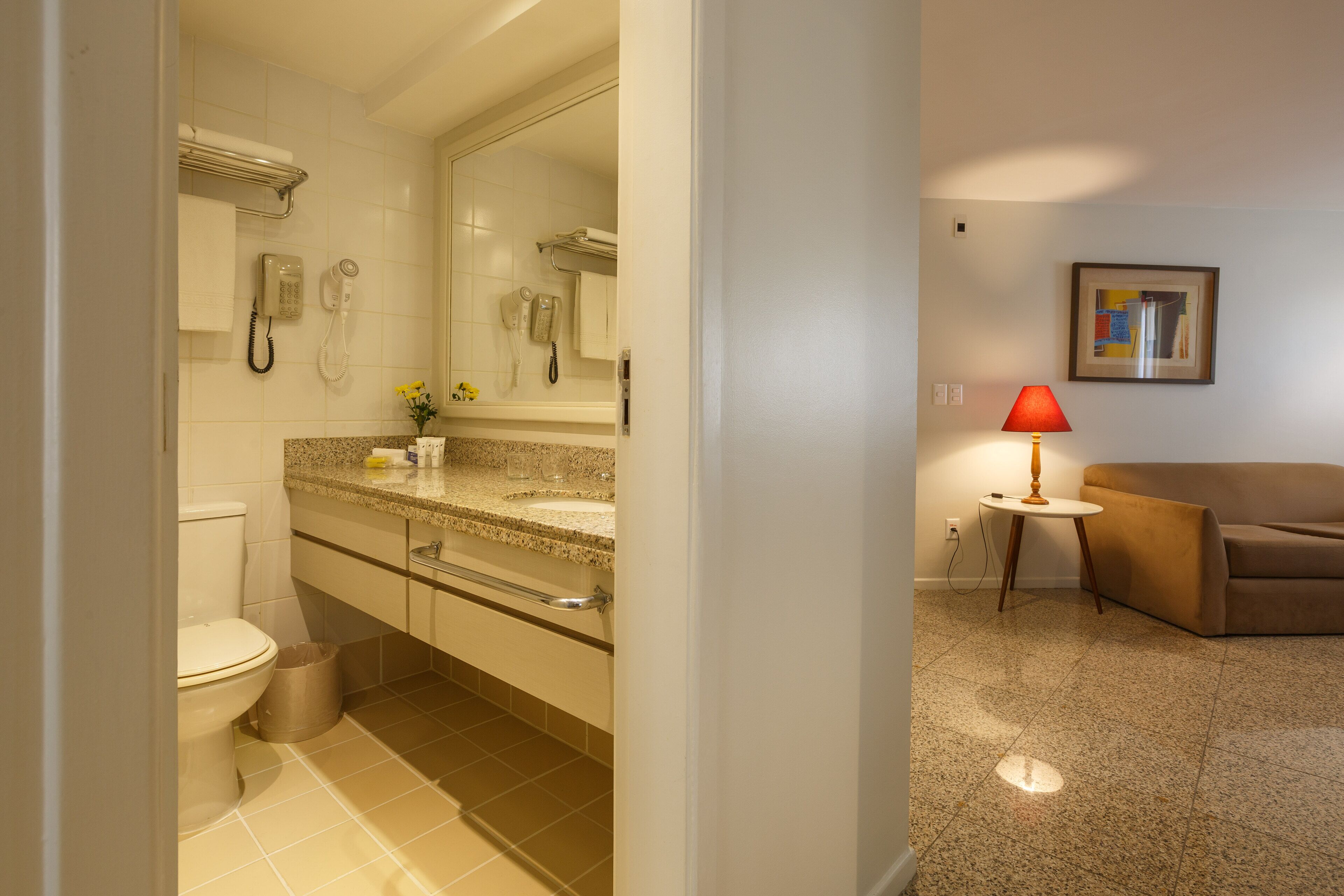 A bathroom with a toilet, sink, and mirror. There is a trash can and a towel rack. A red lamp is on a small table next to a couch.