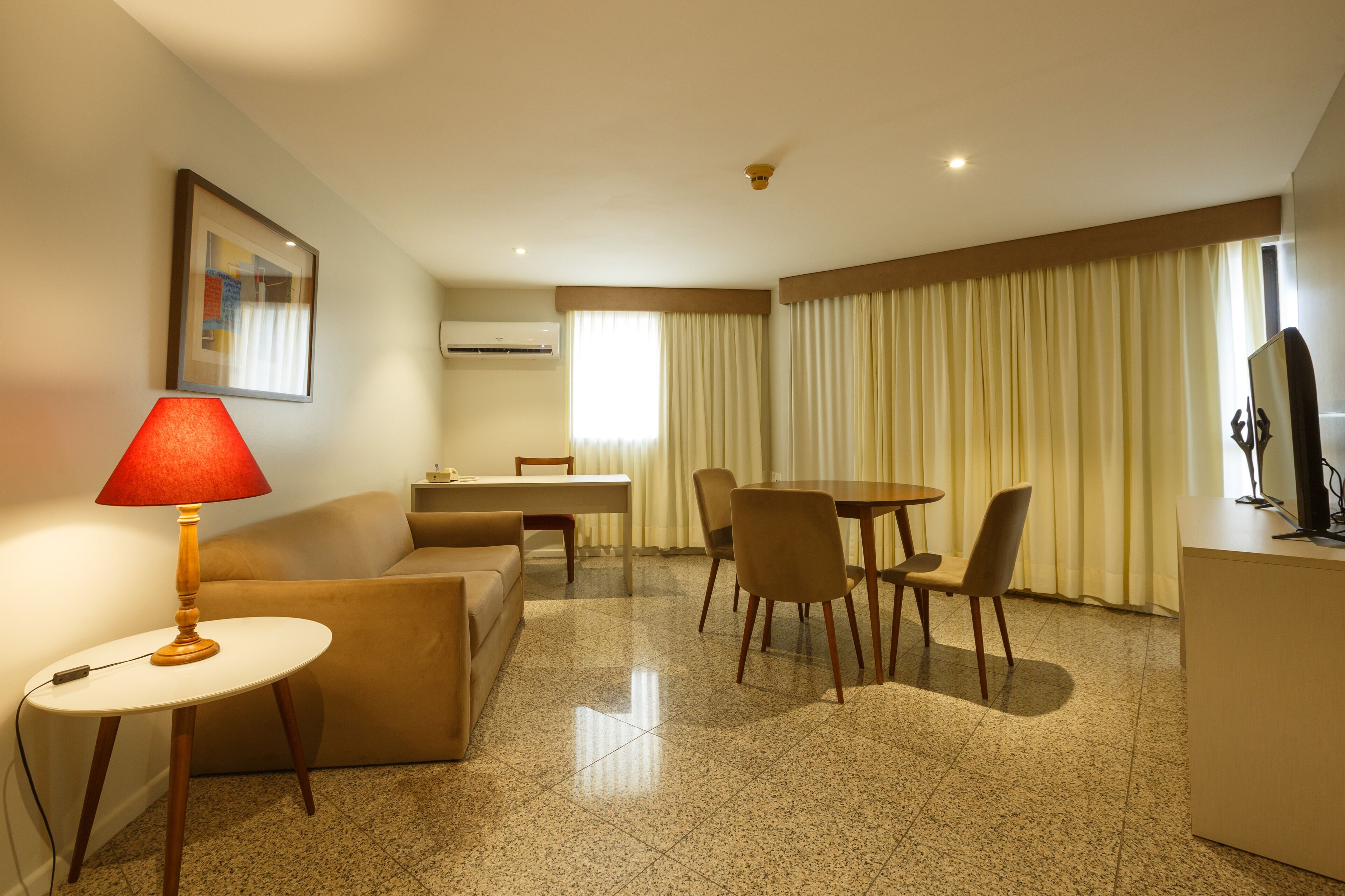 A hotel room with a dining area, a sofa, a table with a lamp, and a TV.