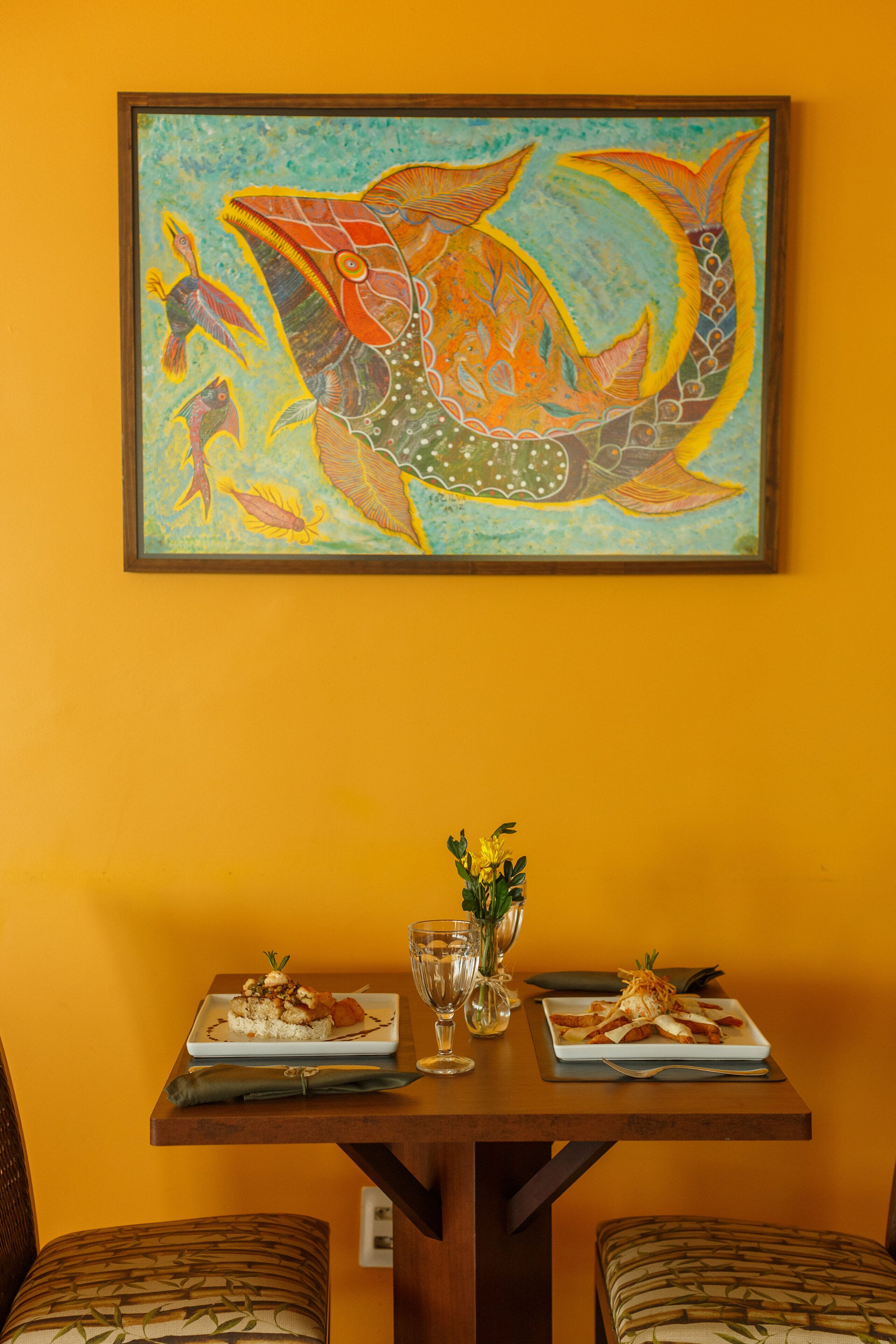 Una mesa de comedor con dos platos de comida, un florero con flores y un cuadro enmarcado de peces en la pared.