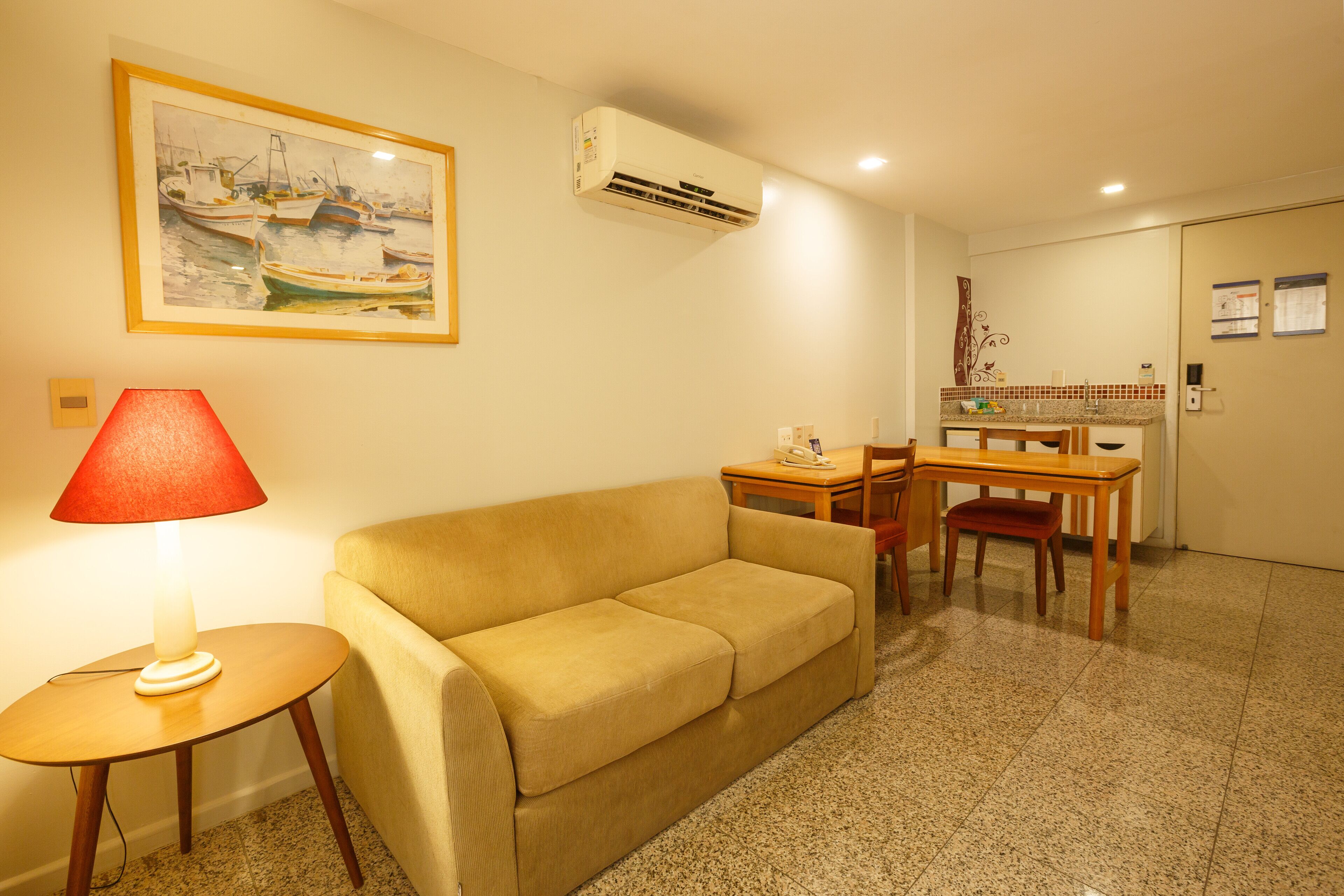 A living room with a sofa, a table with a lamp, a dining table with chairs, an air conditioner, and a painting on the wall.