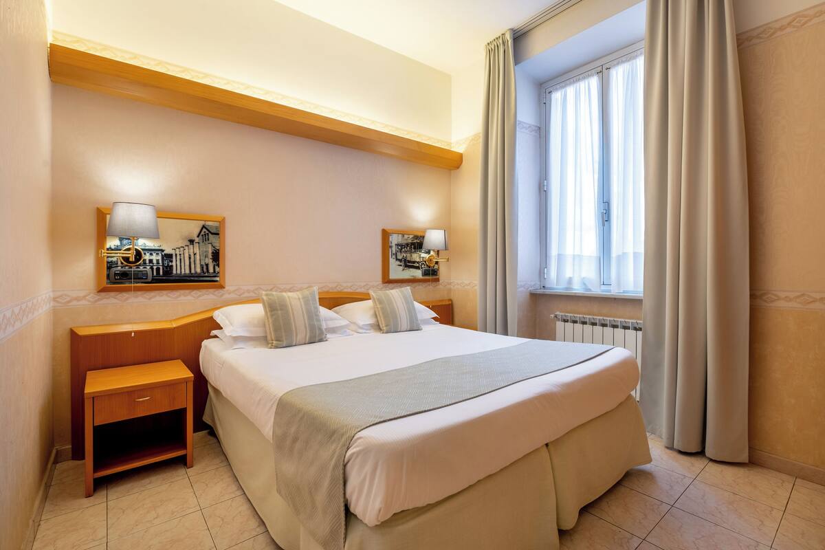 A hotel room with a large bed, two bedside tables, a window with curtains, and wall-mounted lamps.