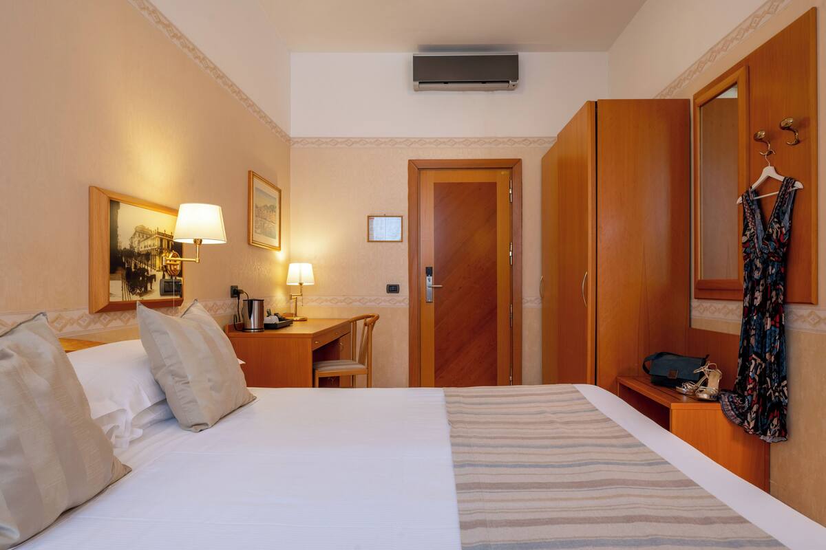 A hotel room with a bed, a desk, a chair, a wardrobe, and a hanging coat.