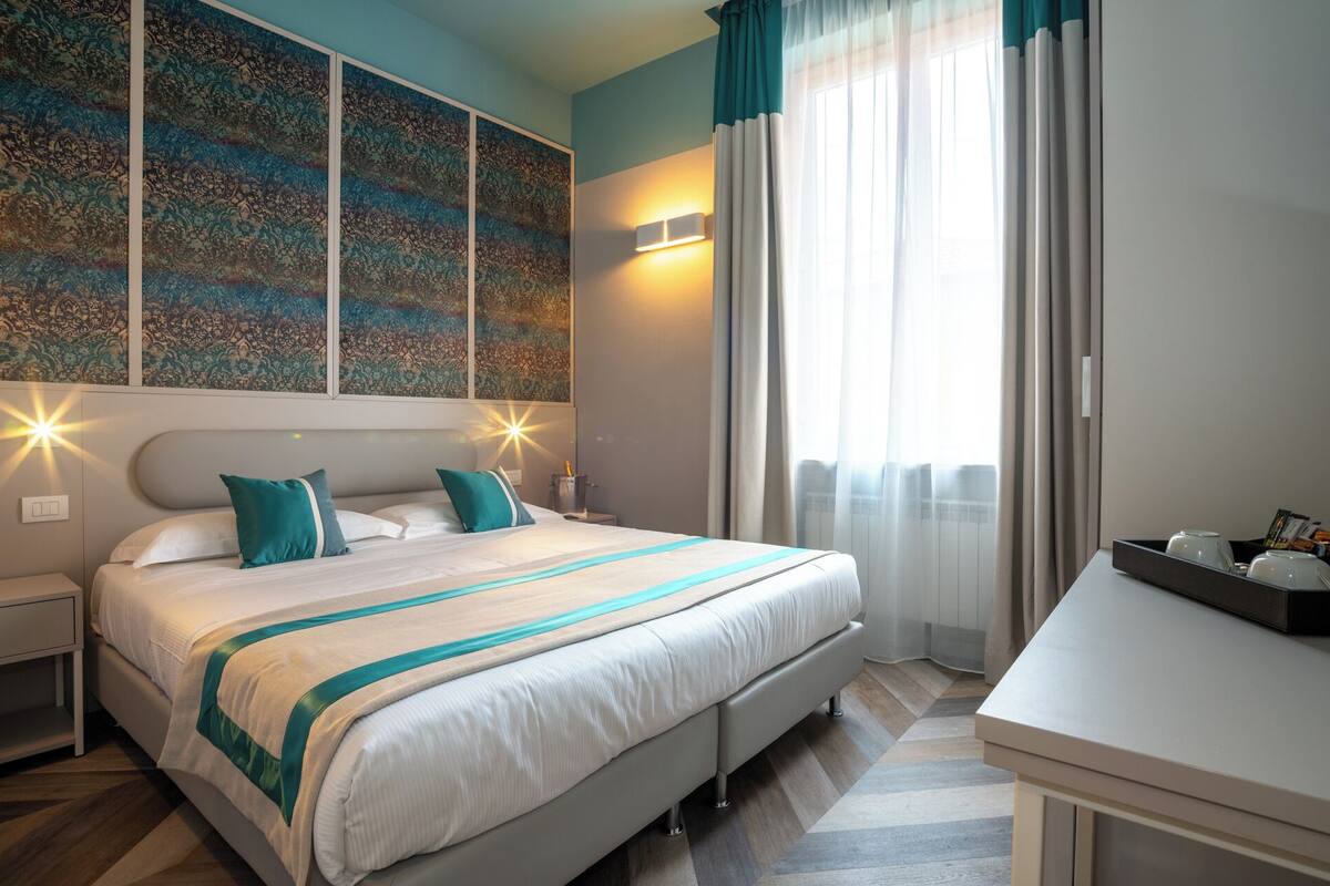 A modern hotel room with a large bed, a desk, and a view of the city through sheer curtains.