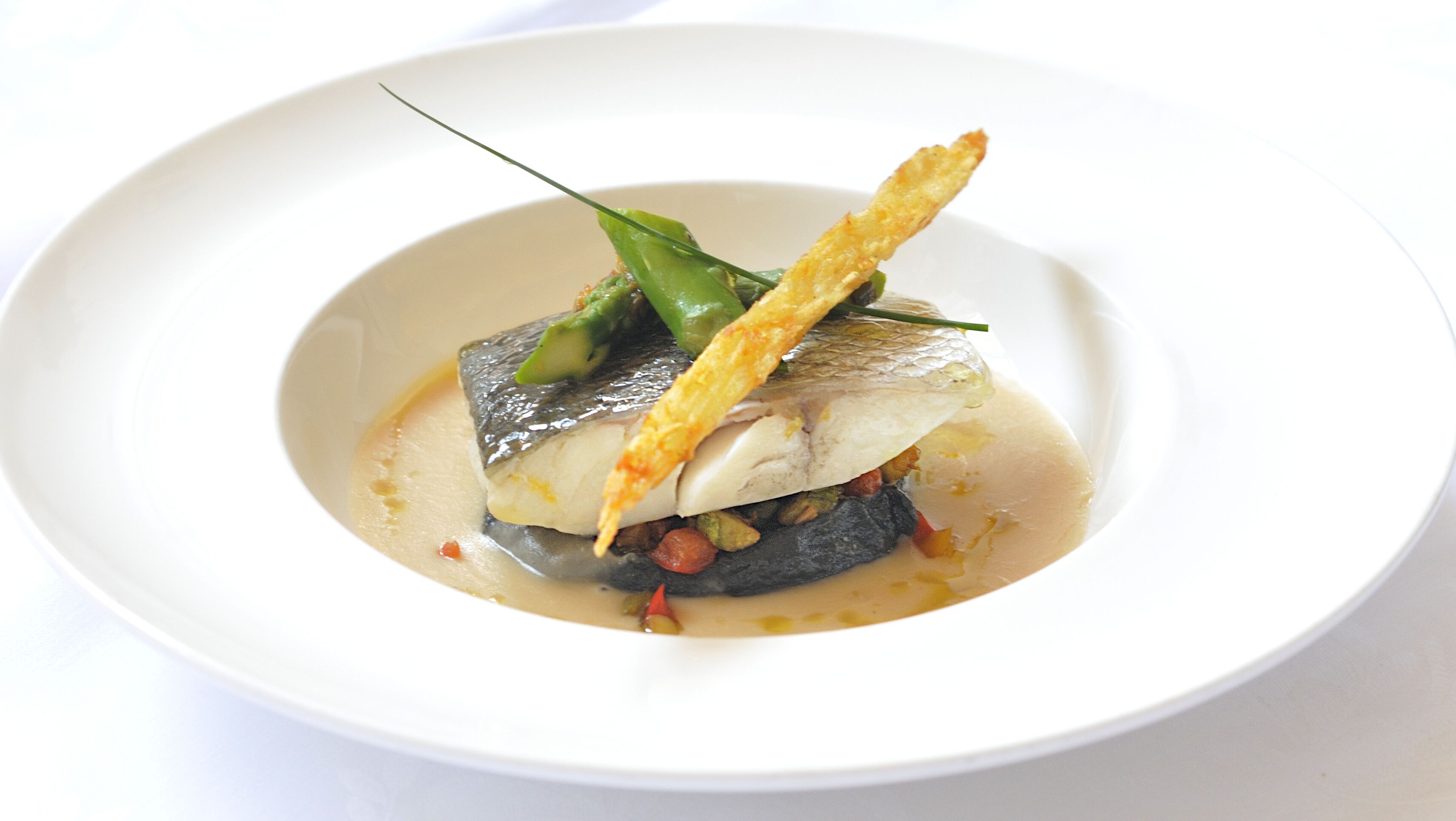 Un plato blanco con un guiso de pescado, verduras verdes y un elemento crujiente.