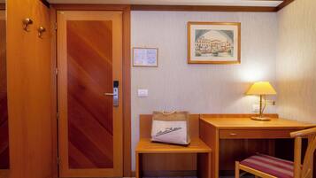 A hotel room with a wooden door, a desk with a lamp, a chair, a small table with a bag, and a framed picture on the wall.