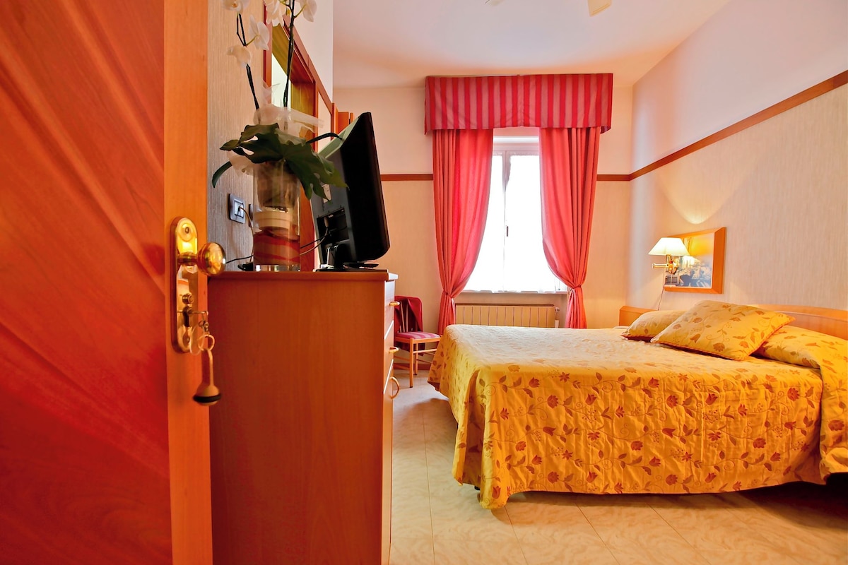 Hotellihuone, jossa on sänky, työpöytä tietokoneella, tuoli, ikkuna verhoilla ja lamppu.