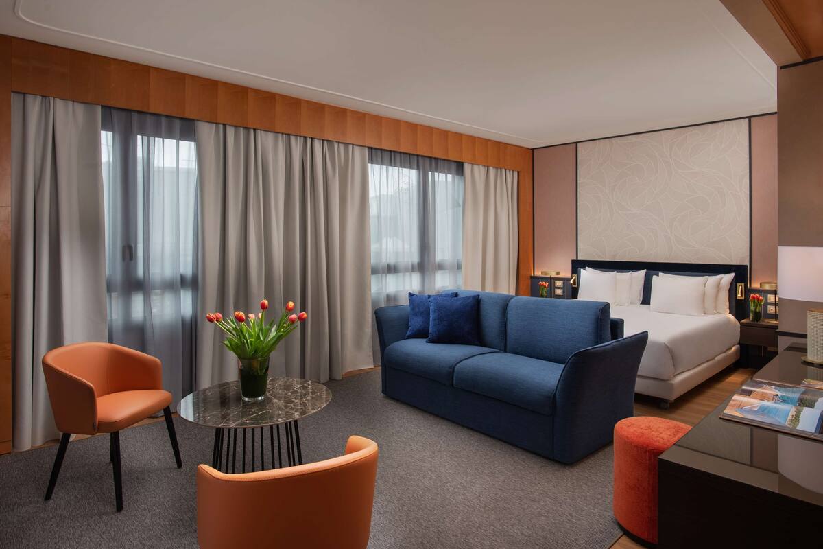 A modern hotel room with a blue sofa, a bed, a round table with a vase of flowers, and a view of the city through large windows.