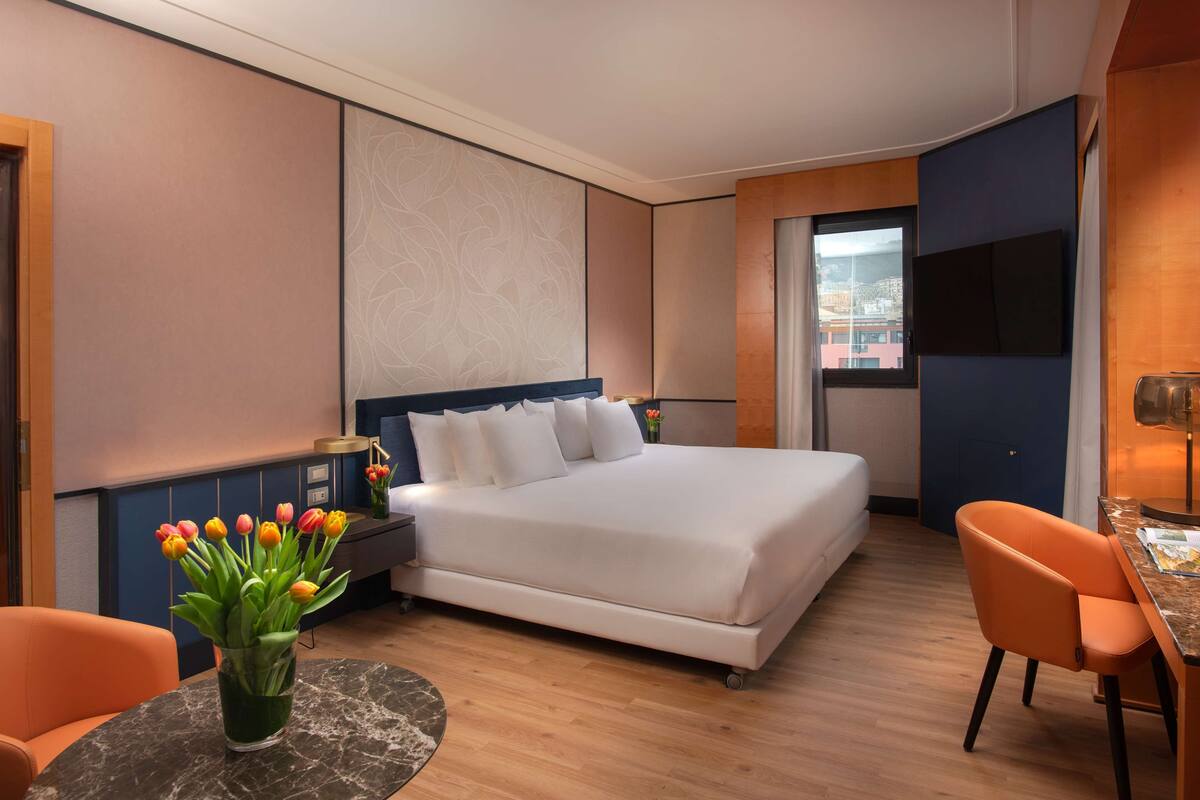A modern hotel room with a large bed, a desk with a chair, a small table with a vase of flowers, and a view of the city through a window.