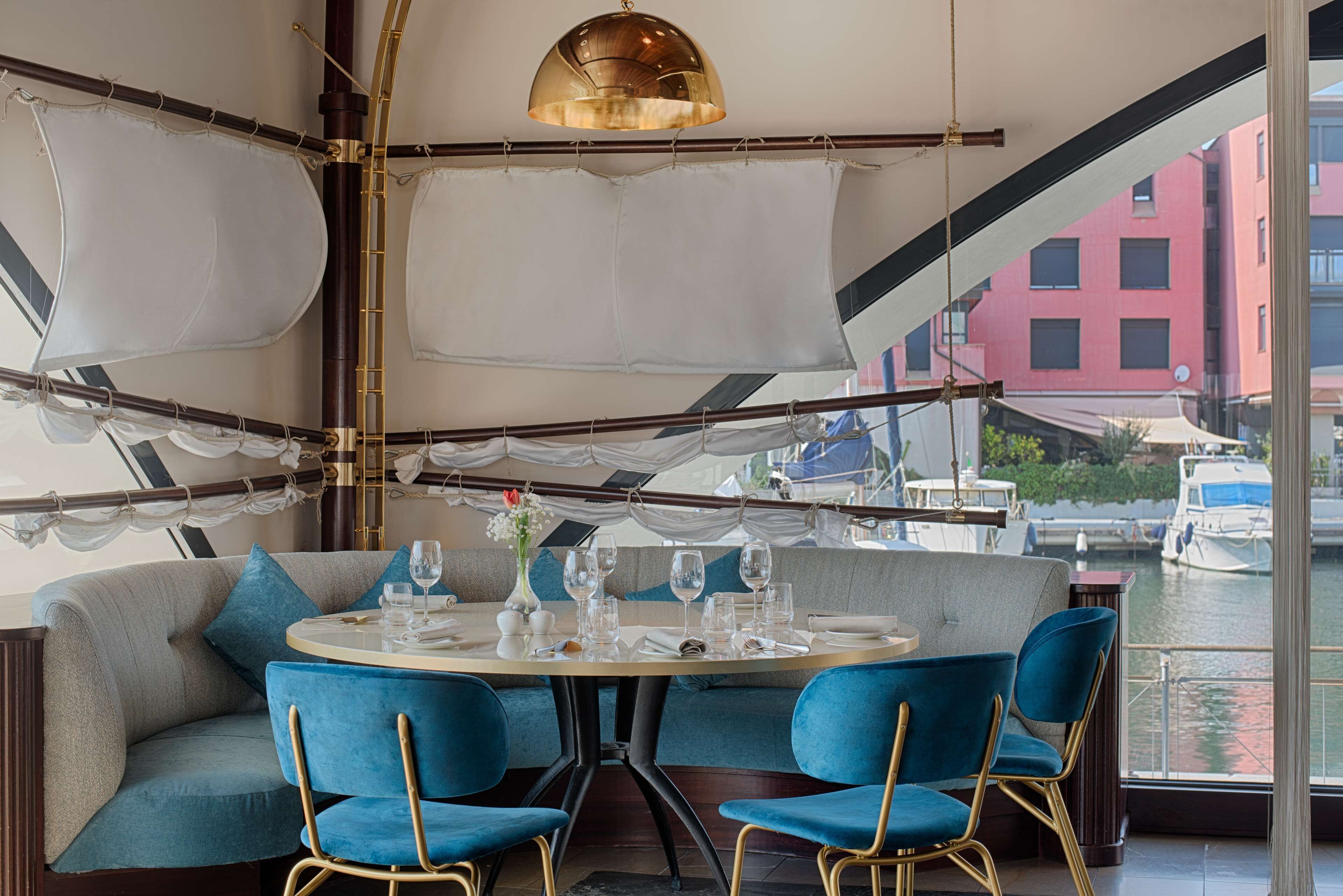 Un coin repas avec une table ronde, des chaises bleues et une vue sur les bateaux à travers de grandes fenêtres.