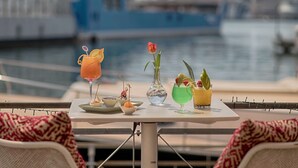 Une table avec diverses boissons et un vase avec une fleur, le tout placé devant un port de plaisance.