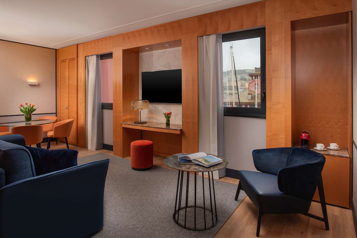 A modern hotel room with a wooden panel wall, a flat-screen TV, a dining area with orange chairs, a red ottoman, and a small round table with a book.