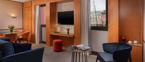 A modern hotel room with a wooden panel wall, a flat-screen TV, a dining area with orange chairs, a red ottoman, and a small round table with a book.