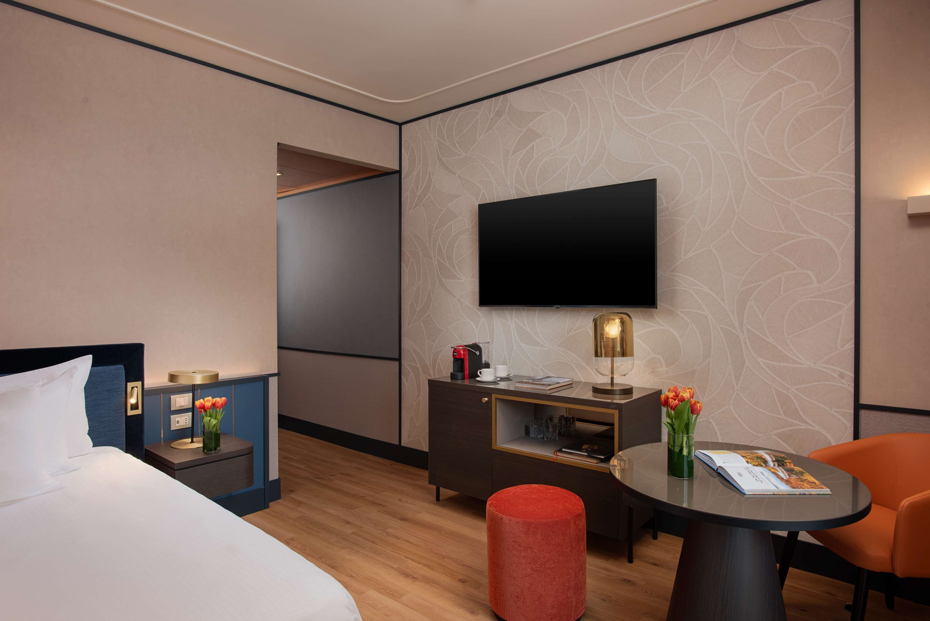 A hotel room with a bed, a desk with a flat-screen TV, a red ottoman, and a small table with a magazine.