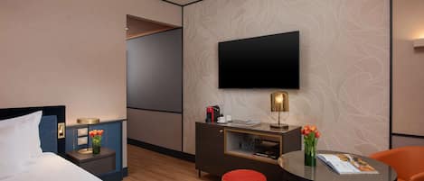A hotel room with a bed, a desk with a flat-screen TV, a red ottoman, and a small table with a magazine.