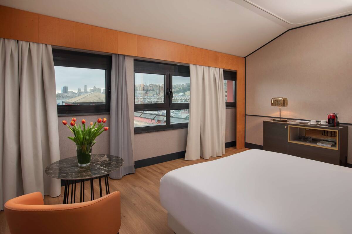 A hotel room with a bed, a desk, a chair, a small table with a vase of flowers, and a view of a cityscape through large windows.