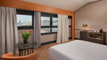 Habitación de hotel con cama, escritorio, silla, una pequeña mesa con un jarrón de flores y vistas a un paisaje urbano a través de grandes ventanales.