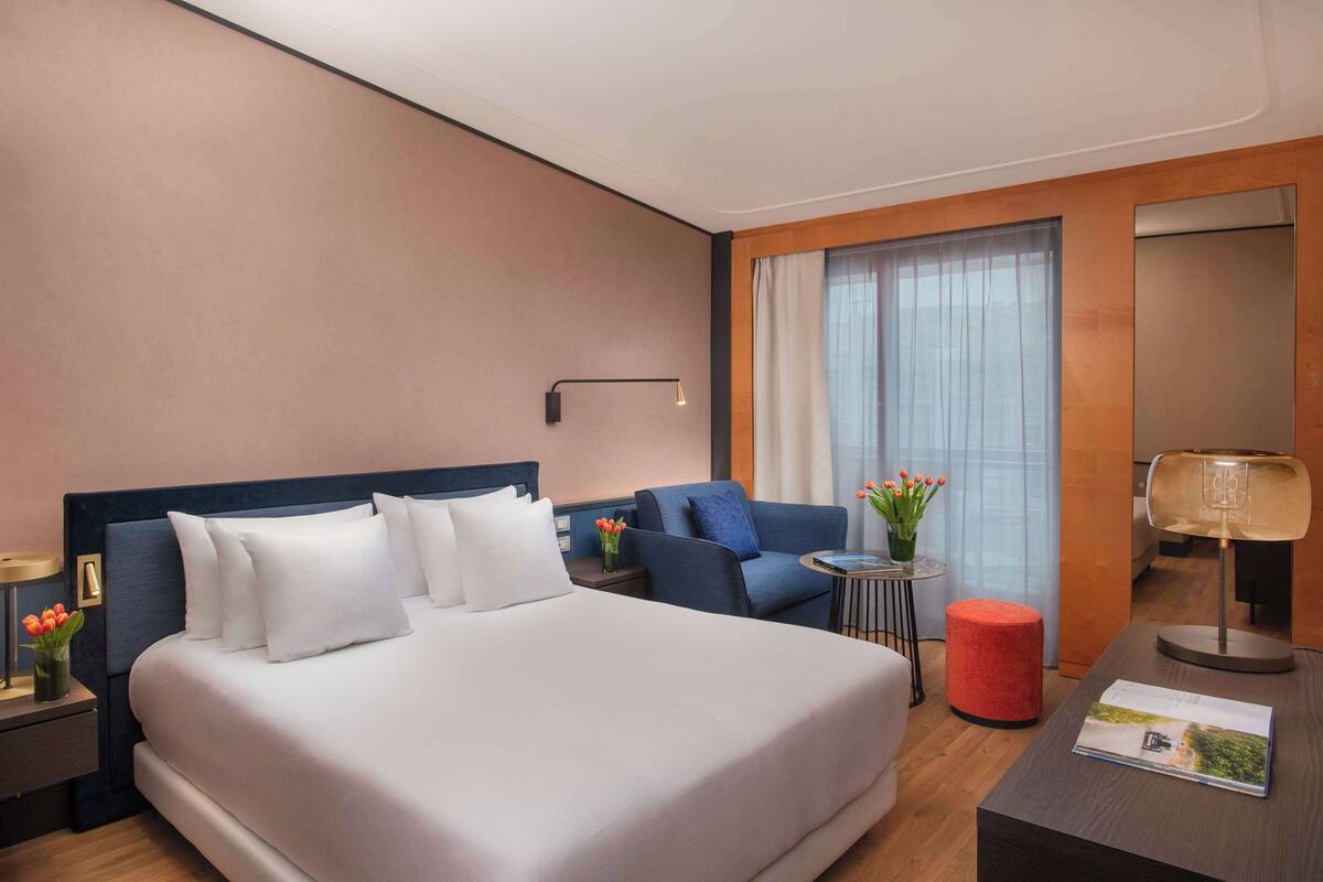 A hotel room with a large bed, a sofa, a small table with a lamp, a red ottoman, and a window with curtains.