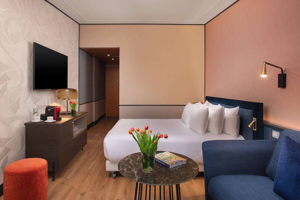 A modern hotel room with a bed, a sofa, a coffee table, a TV, a desk with a lamp, and a vase with flowers.