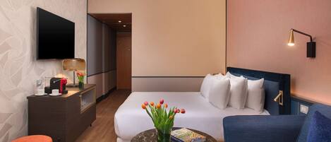 Une chambre d’hôtel moderne comprenant un lit, un canapé, une table basse, un téléviseur, un bureau avec une lampe et un vase avec des fleurs.