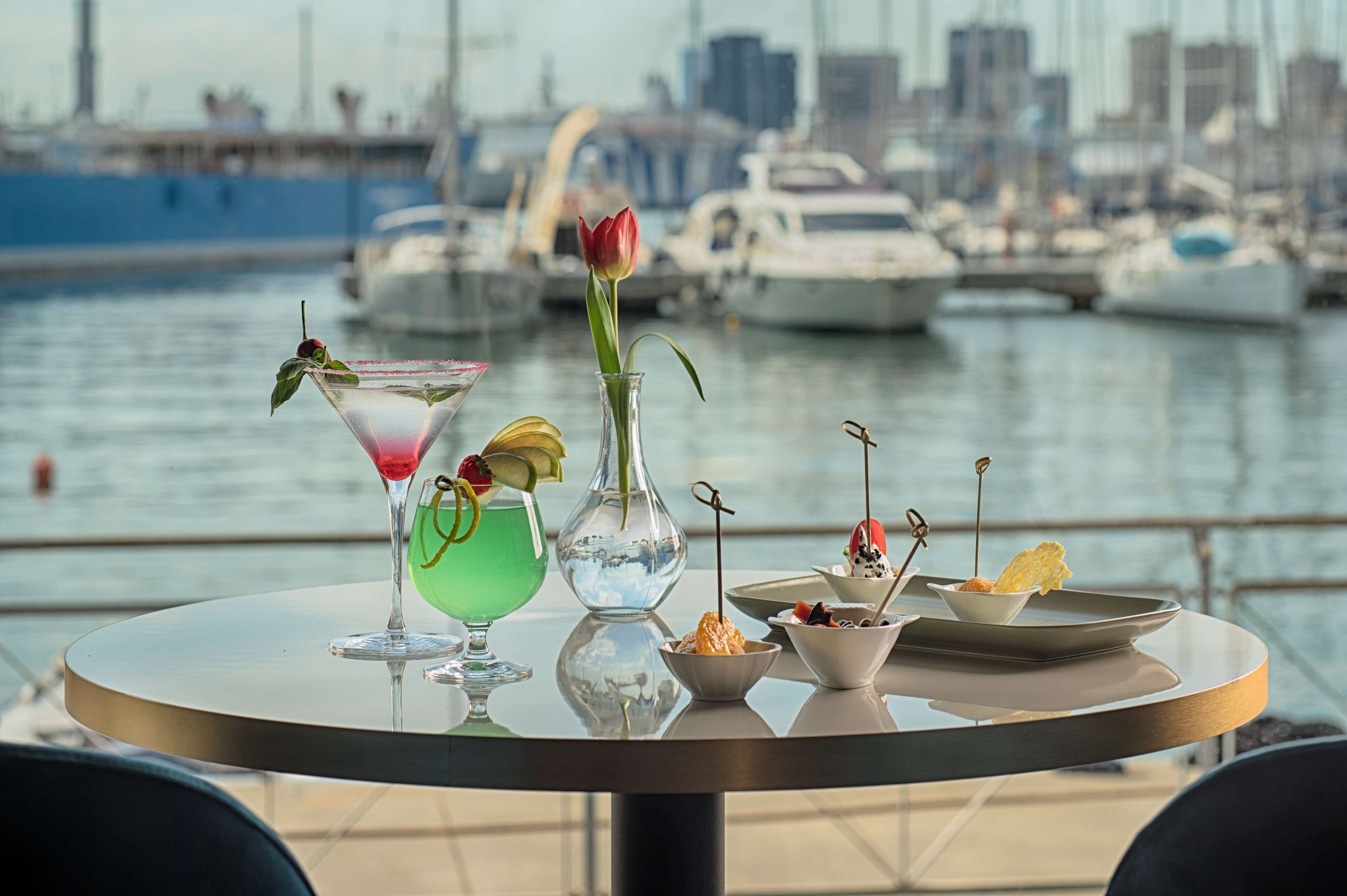 Ein Tisch mit verschiedenen Getränken und Desserts, eine Vase mit einer Blume sowie im Hintergrund der Blick auf eine Stadtmarina.
