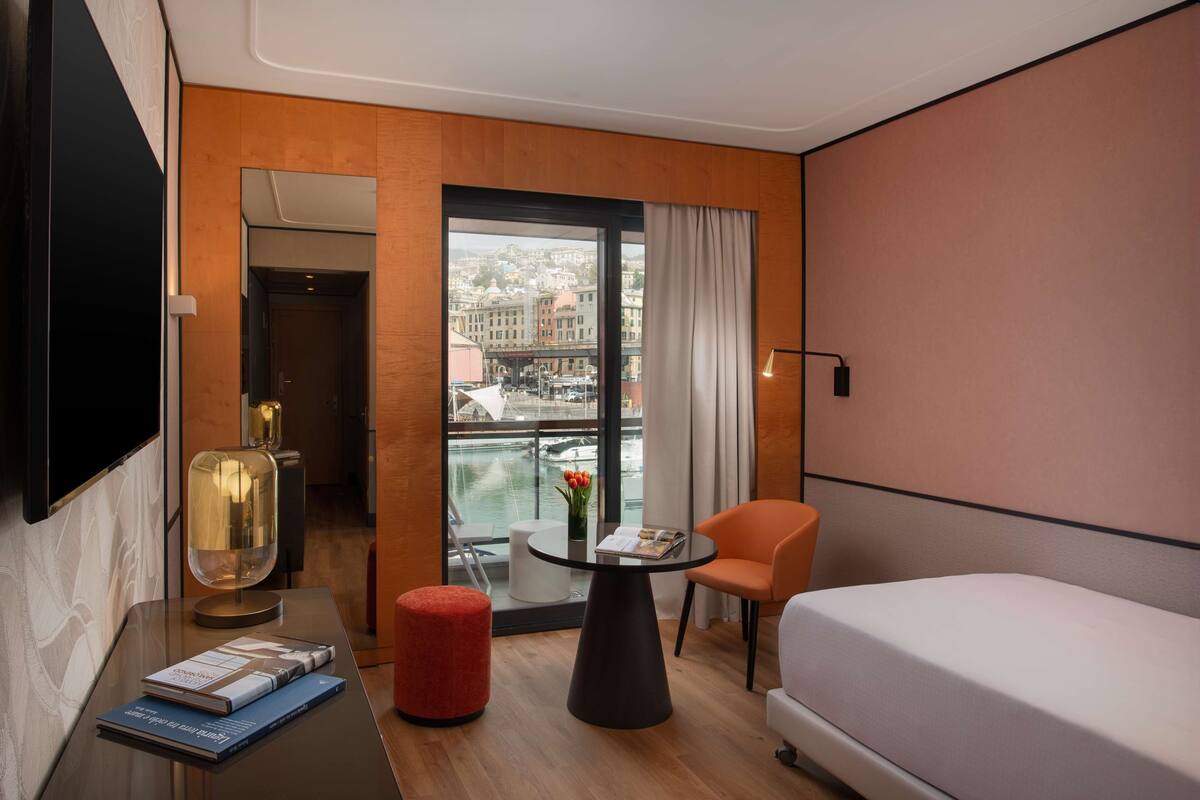 A hotel room with a bed, a small table with a lamp, a red ottoman, a chair, and a view of a cityscape through large windows.