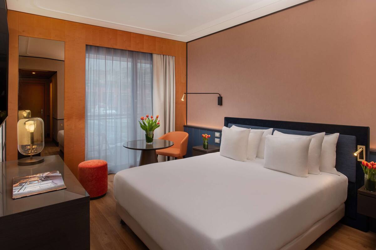 A hotel room with a large bed, a desk with a lamp, a small table with a vase of flowers, and a chair.