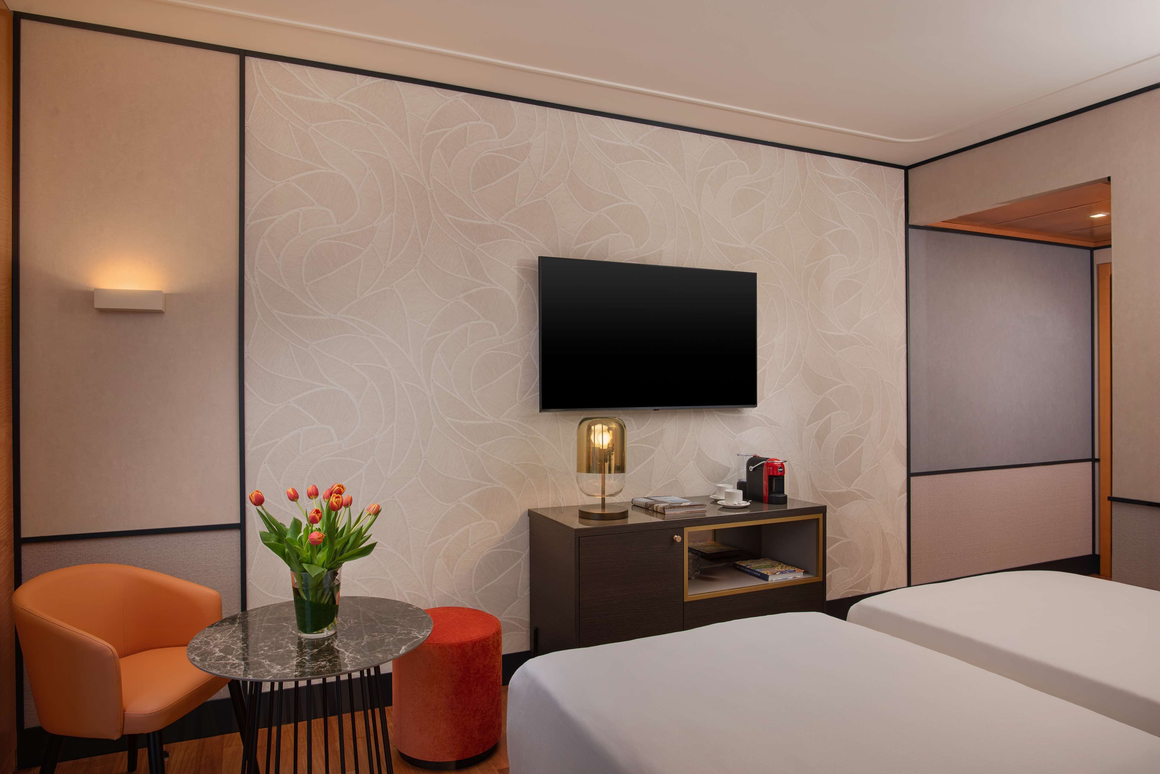 Hotelkamer met een bed, een bureau met een lamp, een televisie, een stoel en een klein tafeltje met een bloevas.