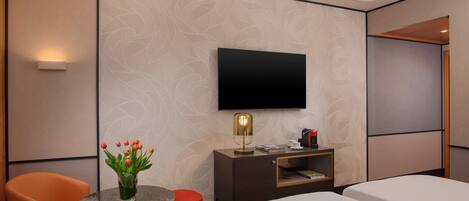 A hotel room with a bed, a desk with a lamp, a TV, a chair, and a small table with a vase of flowers.