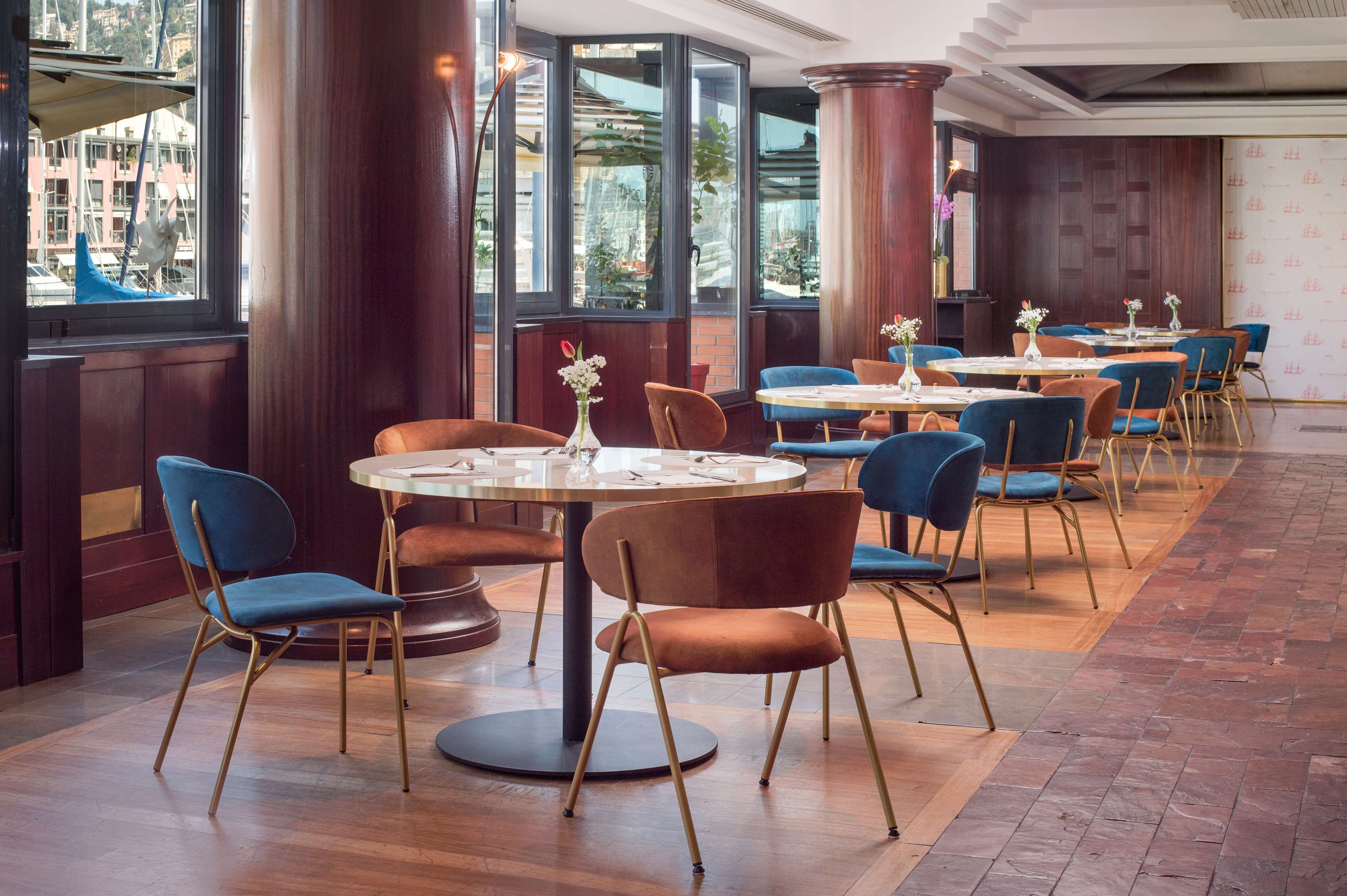 Ein Restaurant mit runden Tischen und blauen Stühlen, großen Fenstern und Holzdielen.