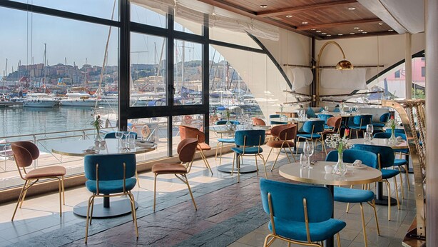 Un restaurant avec des tables et des chaises rondes, de grandes fenêtres donnant sur une marina où sont amarrés des bateaux.