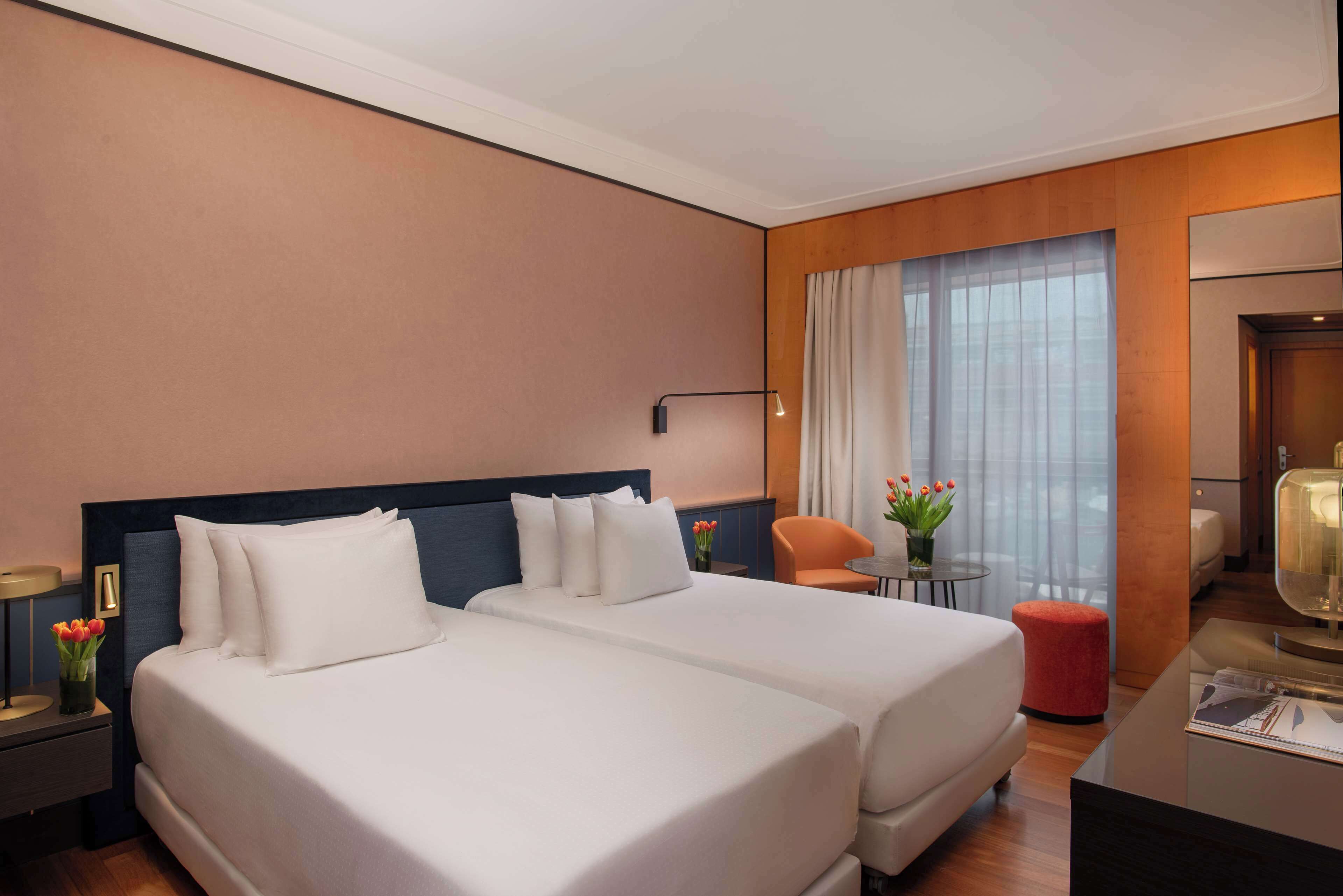 Ein Hotelzimmer mit zwei Betten, einem kleinen Tisch mit Blumenvase, einem Stuhl und einem Schreibtisch mit Lampe.