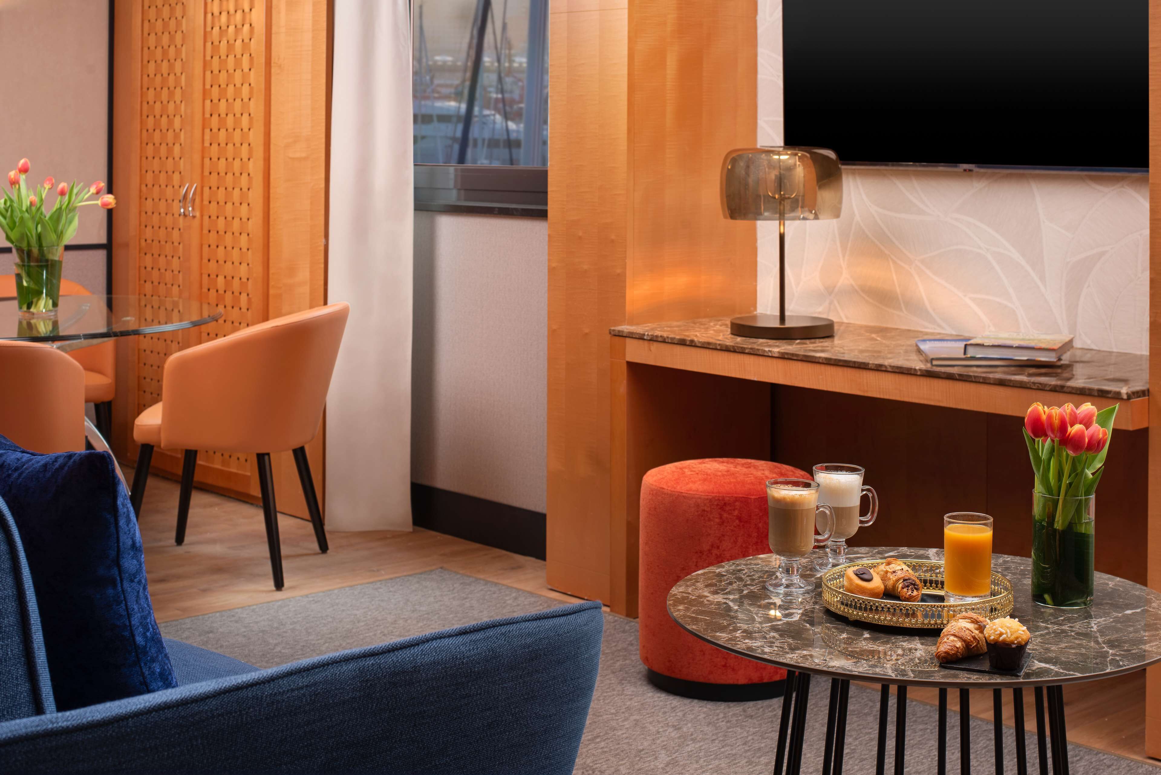 A modern hotel room with a glass dining table, orange chairs, a red ottoman, and a coffee table with breakfast items.