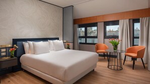 Une chambre d’h ôtel moderne, dotée d’un grand lit, de deux fauteuils, d’une petite table et offrant une vue sur la ville grâce à de grandes fenêtres.