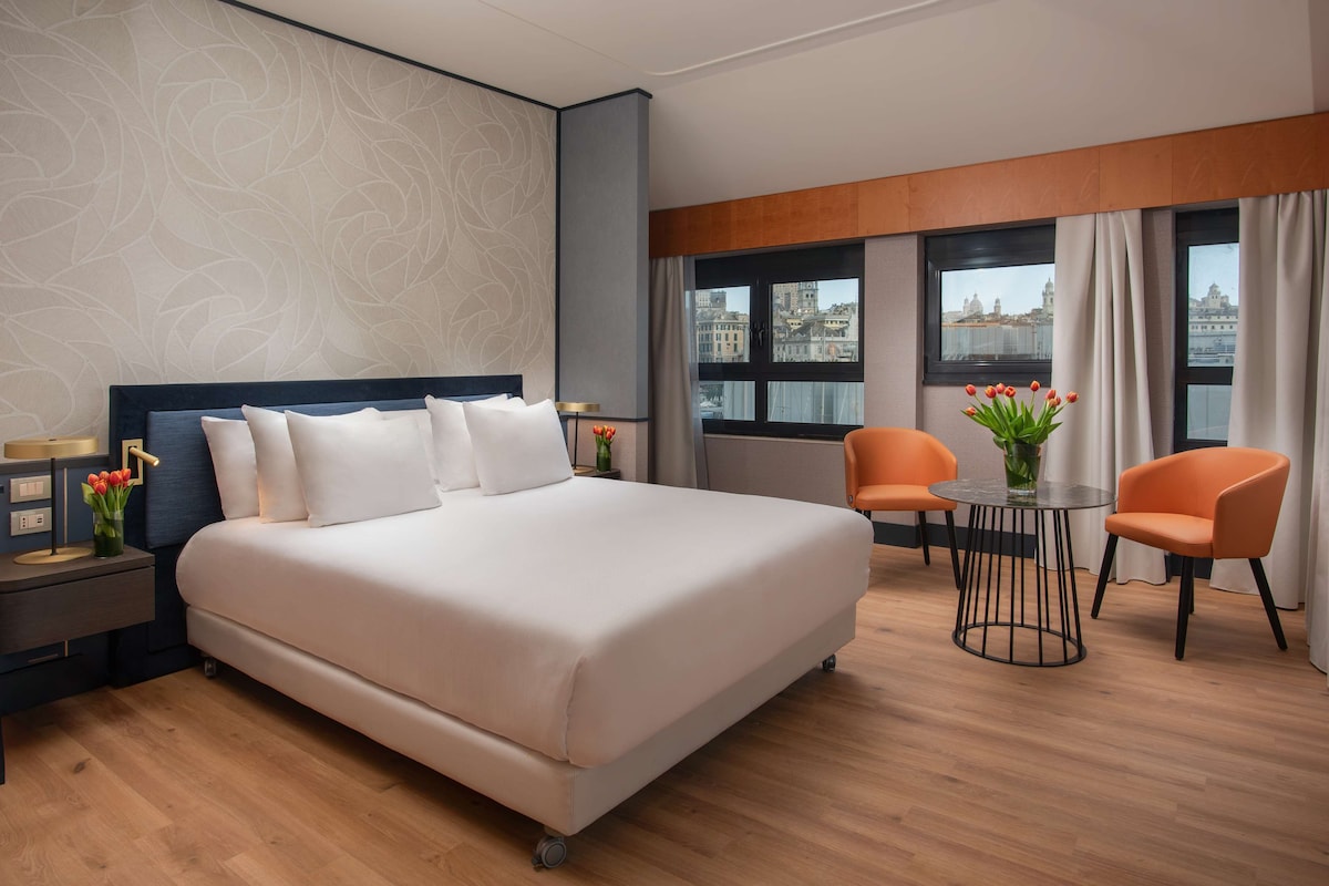 A modern hotel room with a large bed, two chairs, a small table, and a view of the city through large windows.