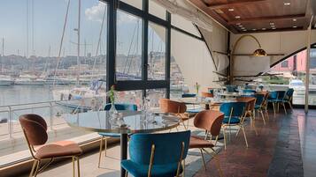 A restaurant with round tables and chairs, large windows overlooking a marina with boats.