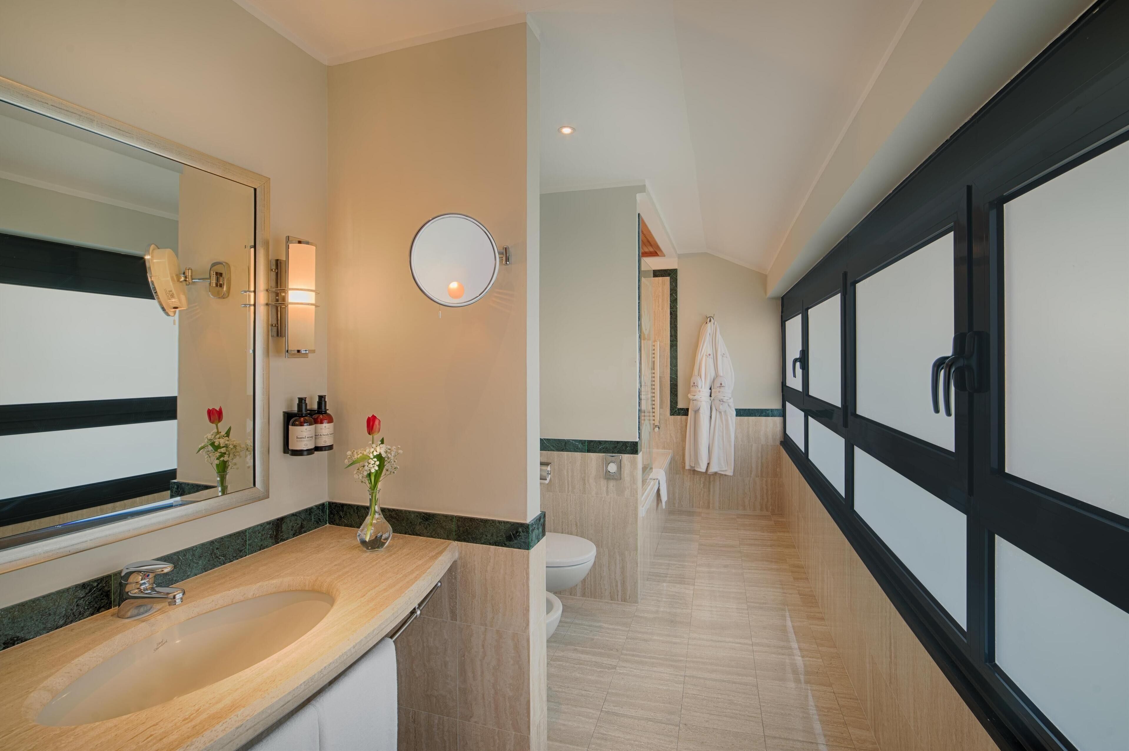 Une salle de bain moderne avec un grand miroir, un lavabo et des toilettes. On y trouve un porte-serviettes et un miroir rond accroché au mur.