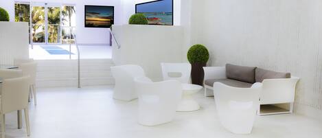 A modern, minimalist lounge area with white furniture, a large flat-screen TV, and a view of palm trees through large windows.