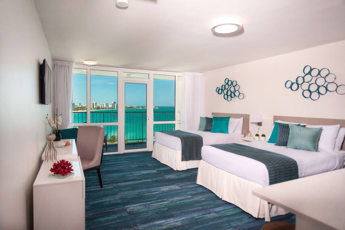 Habitación de hotel con dos camas, un escritorio, una silla y vistas a la ciudad a través de amplios ventanales.