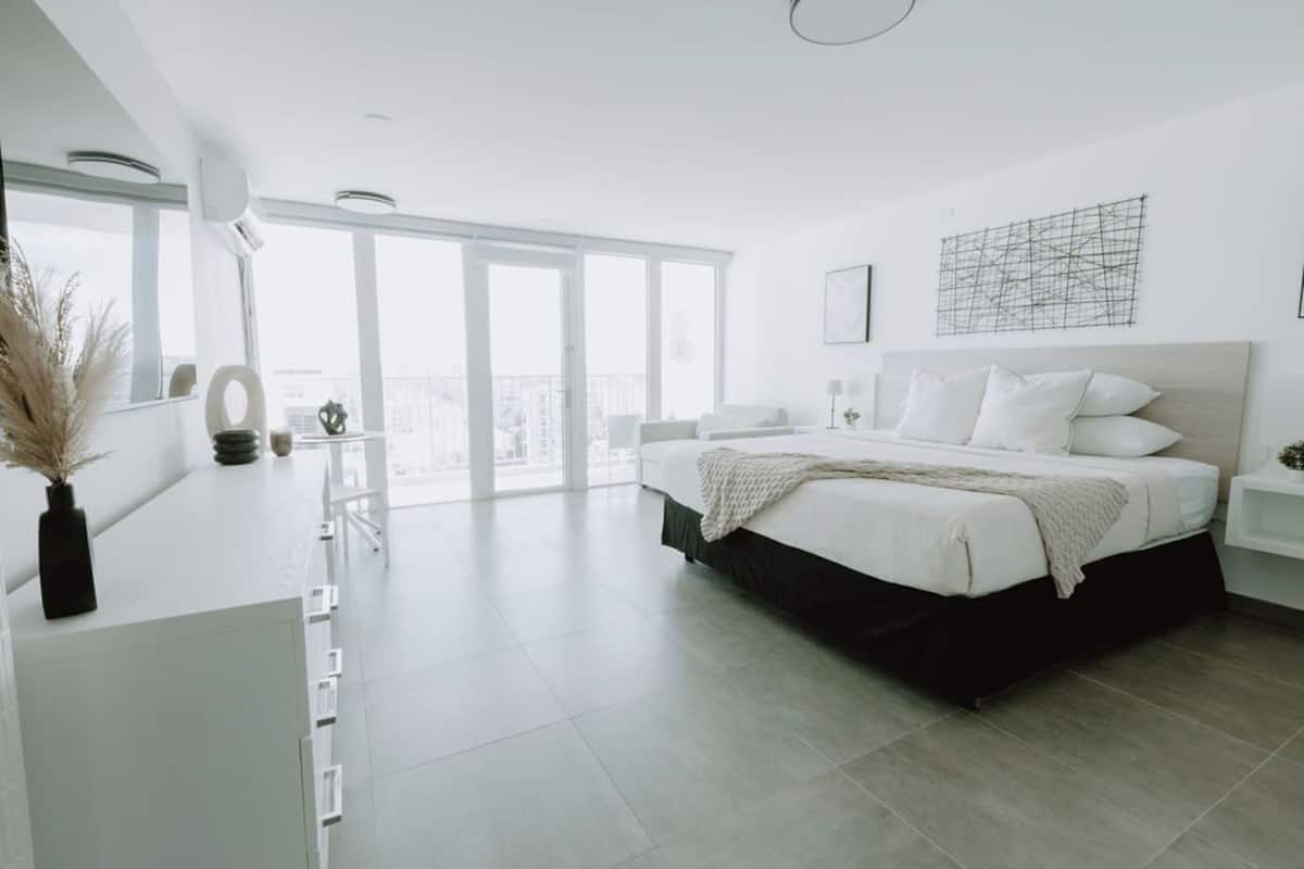 Un dormitorio moderno con una cama grande, un escritorio y vistas de la ciudad a través de amplias ventanas.