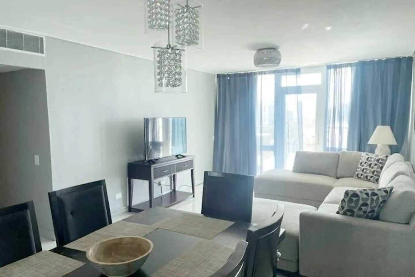 A modern living room with a sofa, a television on a stand, a dining table with chairs, and a large window with curtains.