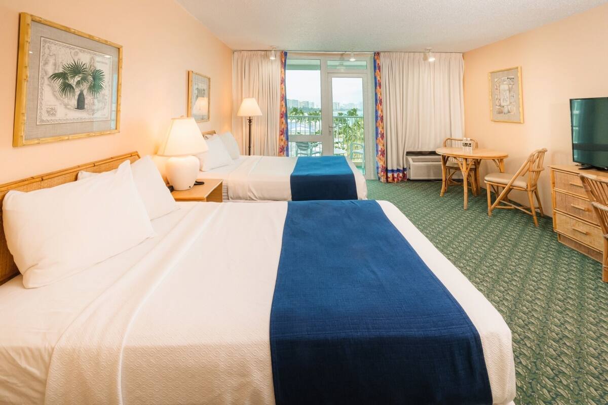 Habitación de hotel con dos camas, un escritorio, una silla, un televisor y un balcón con cortinas.