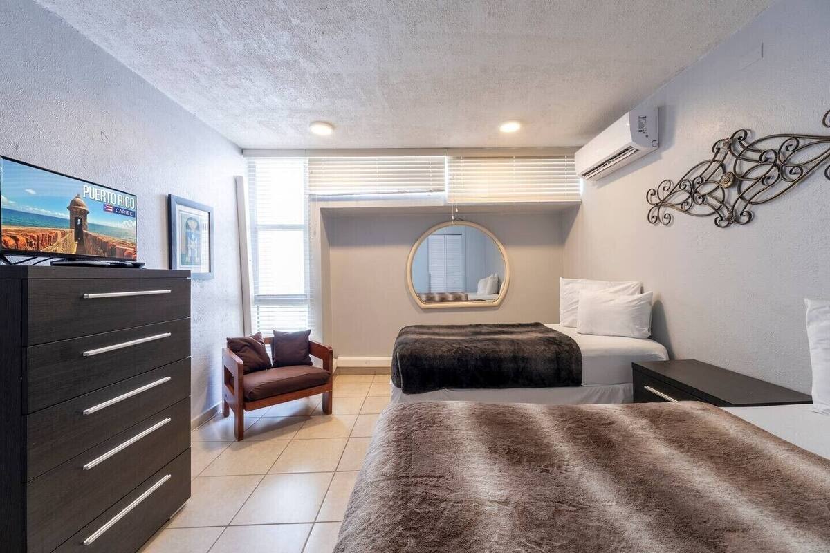 Un dormitorio con cama, cómoda, silla, televisor y espejo adosado a la pared.