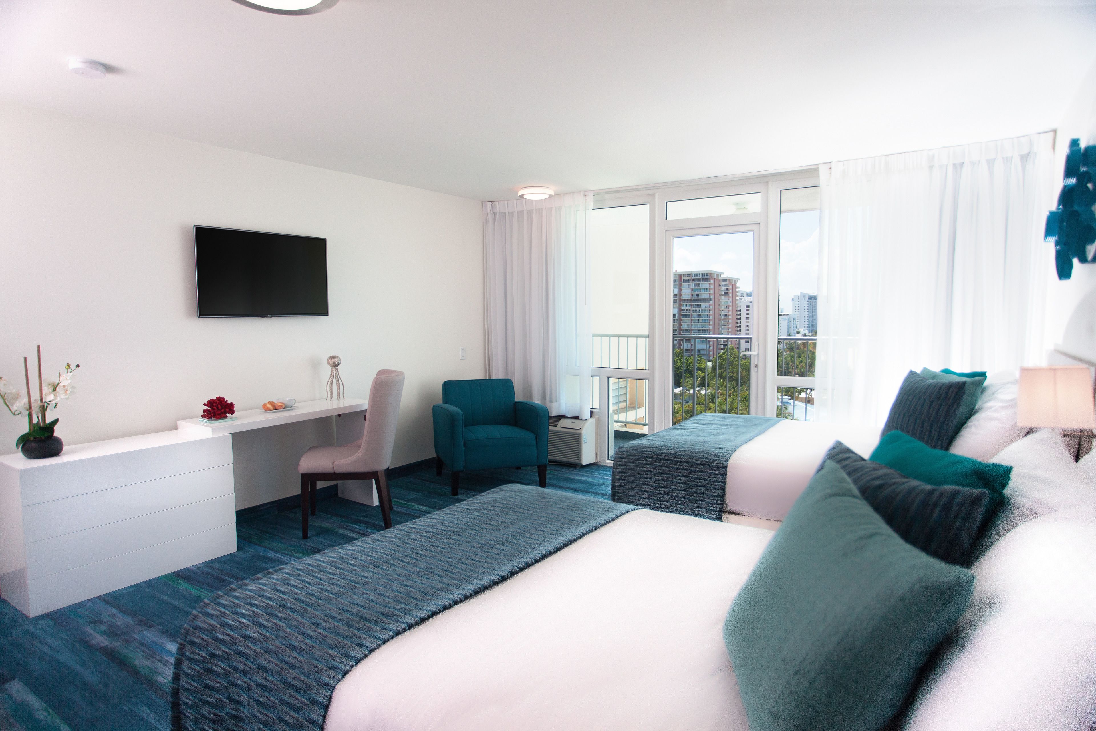 A hotel room with a large bed, a desk, a chair, and a balcony with a city view.