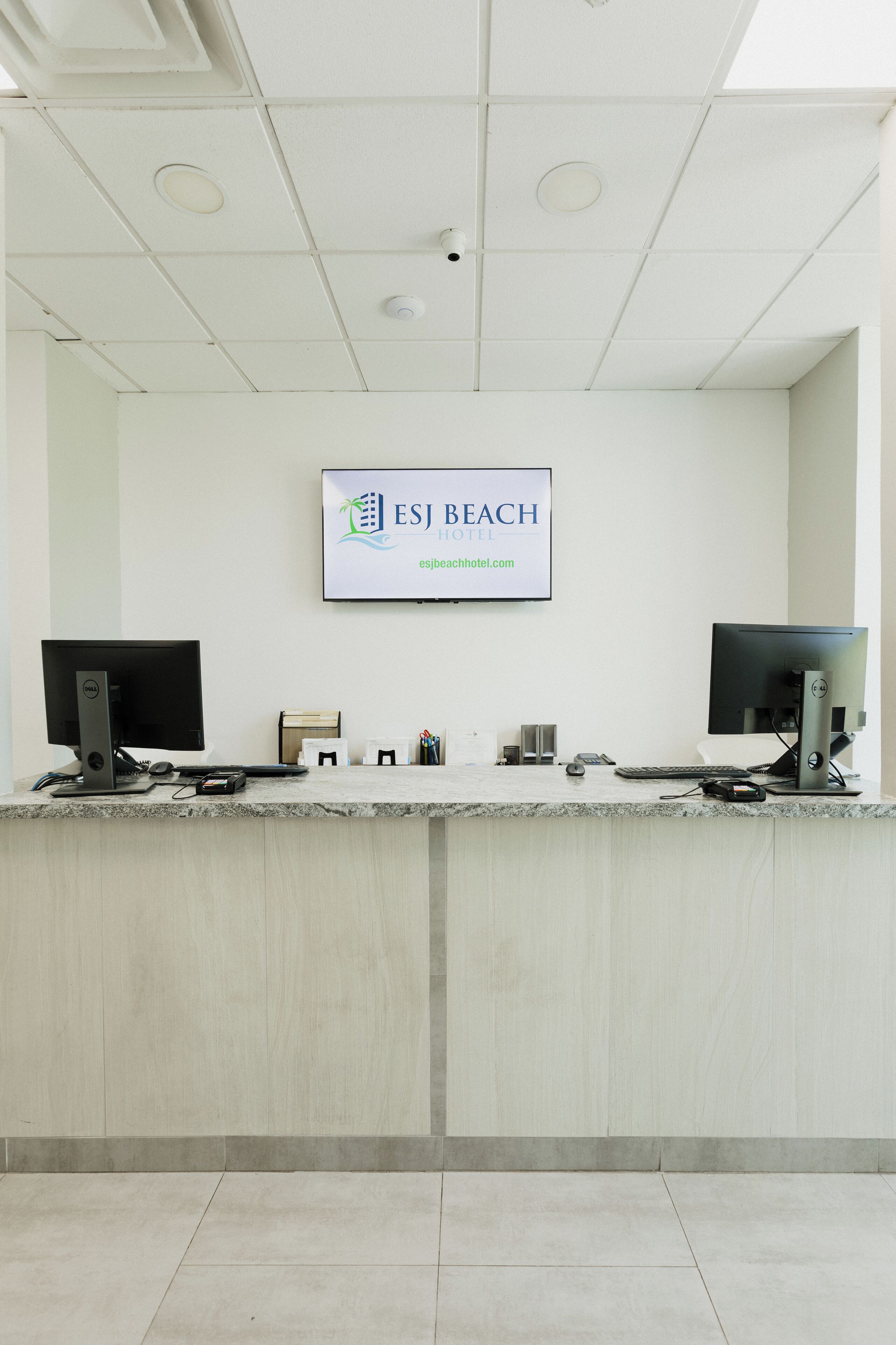 Balcão de recepção de hotel com dois monitores, um computador e uma placa indicando ESJ Beach Hotel.