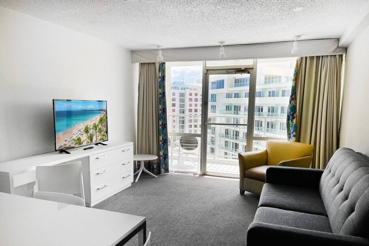 Una habitación de hotel moderna con balcón, televisión de pantalla plana, mesa de comedor, sofá y una silla.