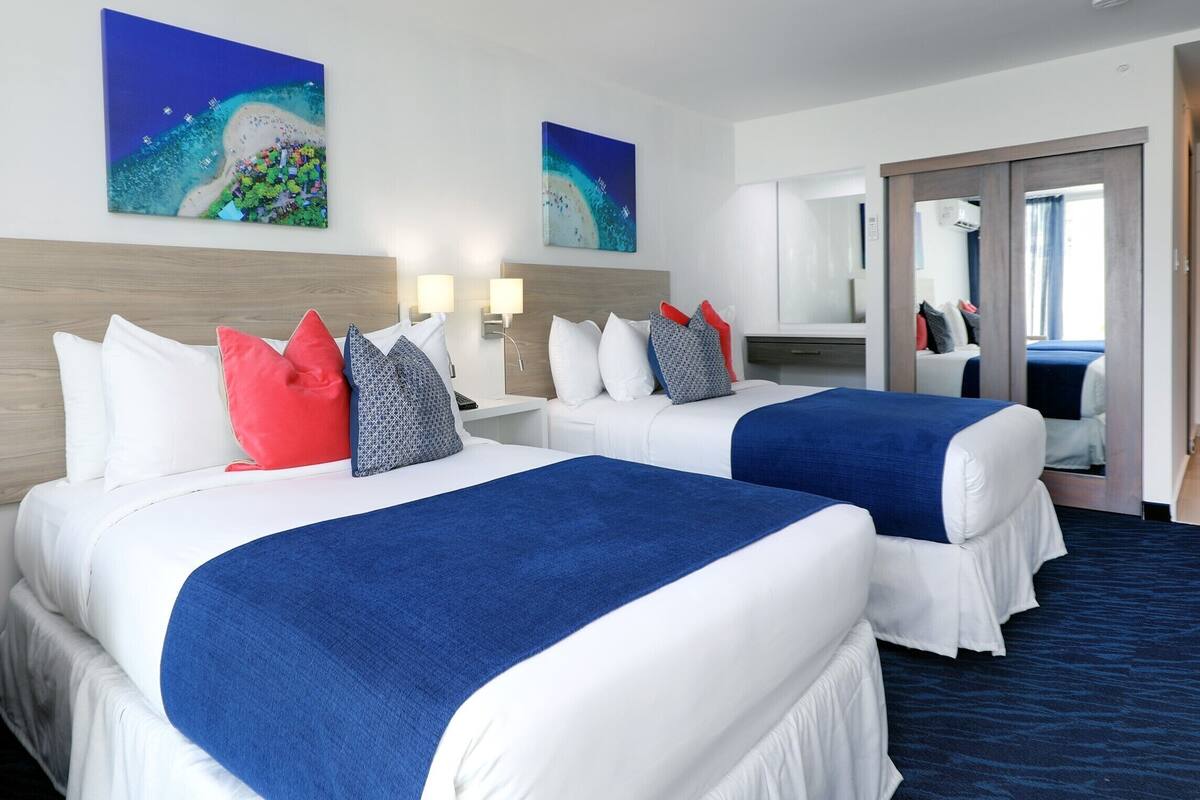 Habitación de hotel con dos camas, cada una con ropa blanca y mantas azules. Hay dos lámparas de pared a cada lado de las camas. La habitación tiene un espejo grande en la pared derecha y una puerta que conduce a otra habitación.