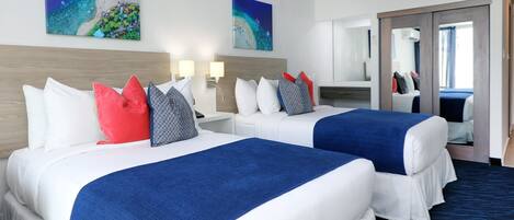 A hotel room with two beds, each with white bedding and blue throw blankets. There are two wall-mounted lamps on either side of the beds. The room has a large mirror on the right wall and a door leading to another room.