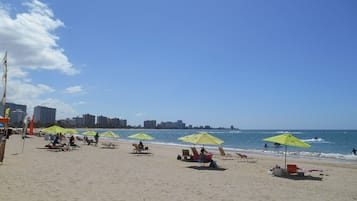 Una playa con filas de sillas y sombrillas, un cielo despejado y un horizonte urbano al fondo.