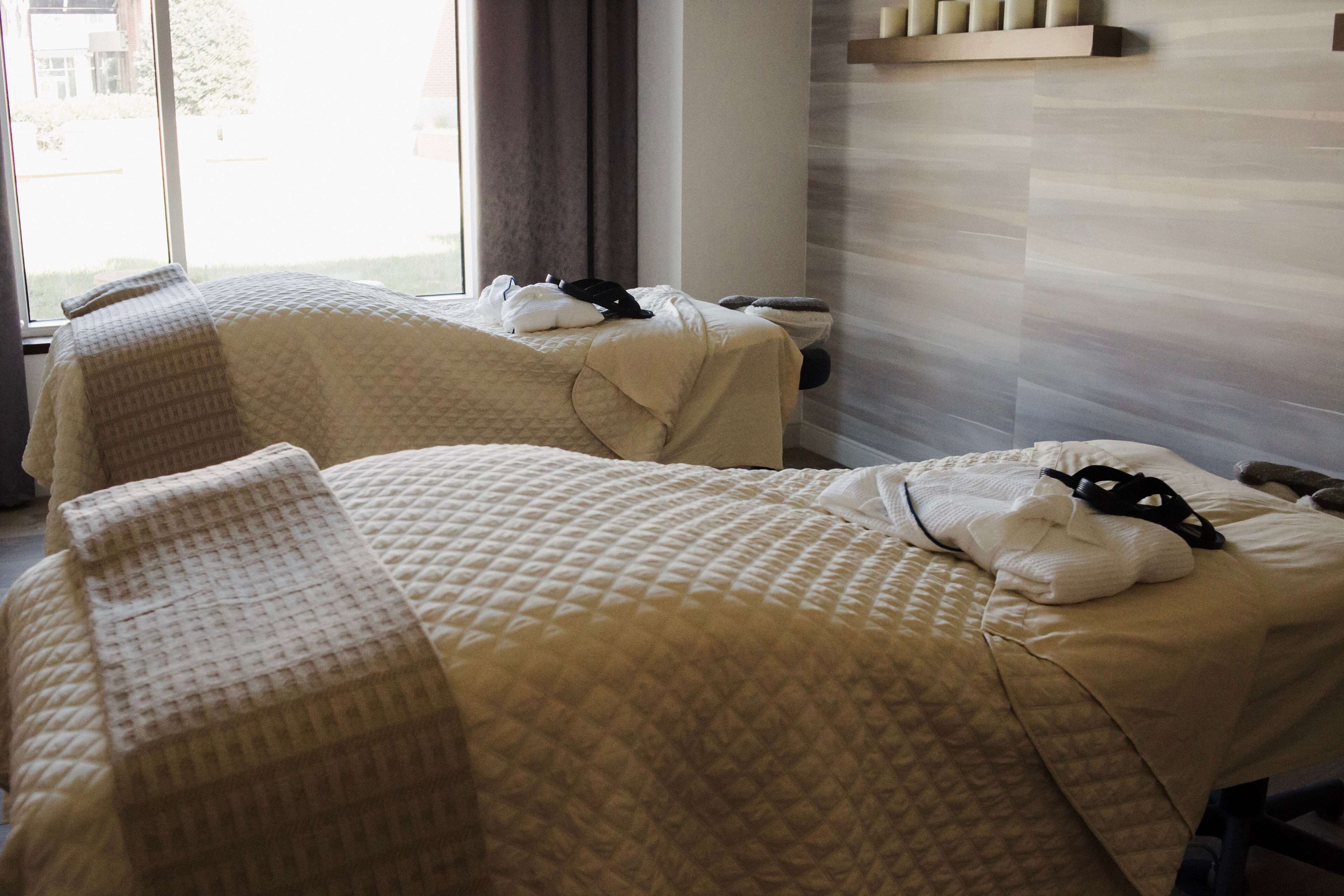 A hotel room with two beds, each with a quilted bedspread and pillows. There is a window with curtains, a wall-mounted shelf with toiletries, and a view of the outdoors.